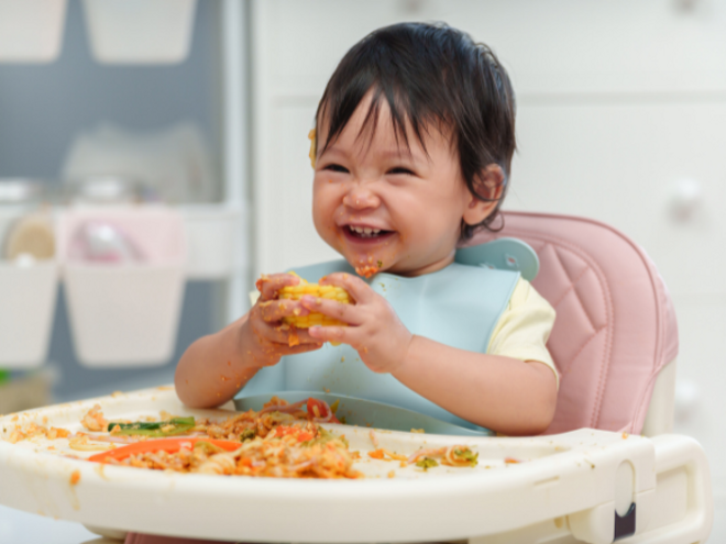 Toddler girl holding corn on a messy highchair
